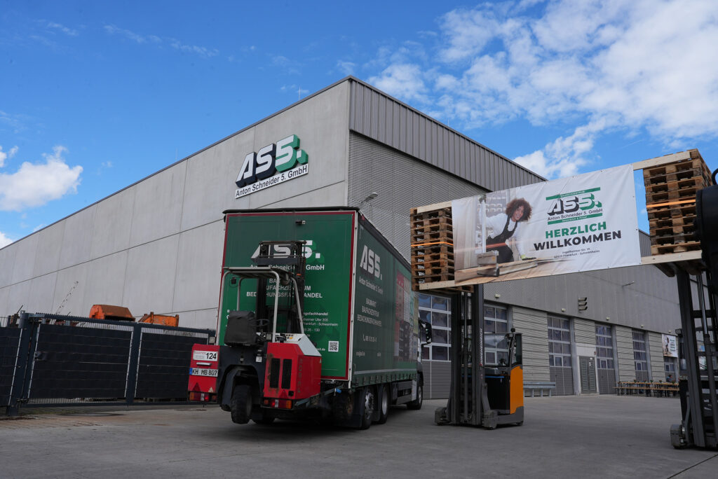 Ein Lkw mit grüner Ladefläche steht vor dem modernen Firmengebäude der Anton Schneider 5. GmbH. Über dem Eingang hängt ein Willkommensbanner mit dem Unternehmenslogo. Im Hintergrund sind Gabelstapler und Paletten sichtbar, während der Himmel teilweise bewölkt ist.